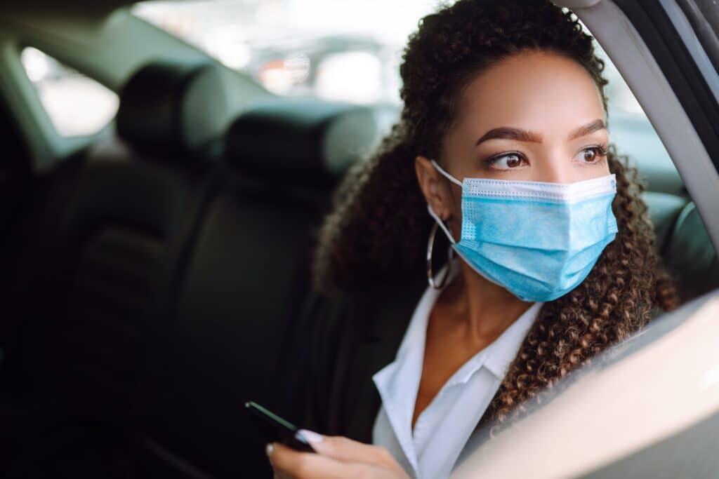 Woman in a sterile mask in taxi car on a back seat looking out of window checking her cell phone.
