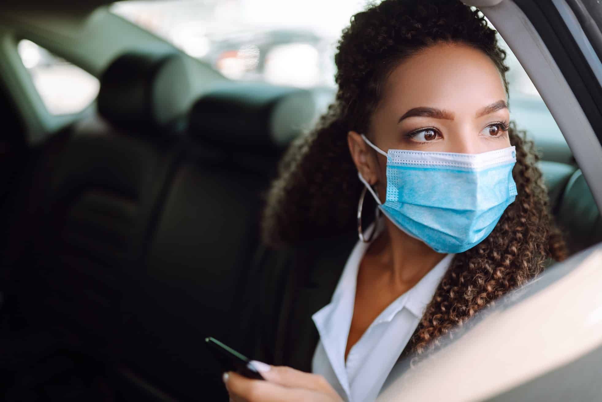 Woman in a sterile mask in taxi car on a back seat looking out of window checking her cell phone.