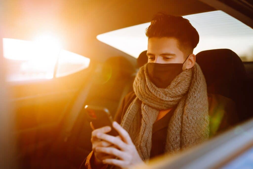 Young man in protective sterile mask in taxi car on a back seat checking her cell phone.